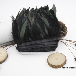 May include: Black feather trim with a green iridescent sheen. The trim is folded and tied with a black ribbon. The trim is on a white surface with wooden accents.