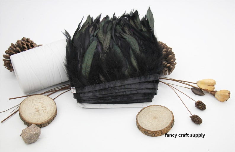 May include: Black feather trim with a green iridescent sheen. The trim is folded and tied with a black ribbon. The trim is on a white surface with wooden accents.