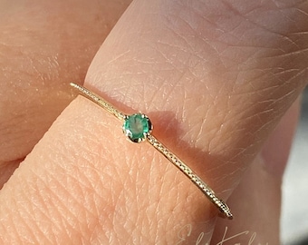 Puede incluir: Un delicado anillo de oro con una pequeña piedra preciosa verde redonda engastada en el centro. El anillo es fino y tiene una banda texturizada.