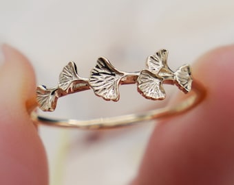 Puede incluir: Un delicado anillo de oro con un diseño de cinco hojas de ginkgo, cada una con intrincados detalles. Las hojas están conectadas por una banda fina, creando un aspecto fluido y elegante.