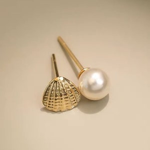 Peut inclure: Deux boucles d'oreilles dorées. L'une est en forme de coquillage texturé. L'autre est une tige dorée avec une grosse perle blanche ronde. Les boucles d'oreilles sont présentées sur une surface beige clair.