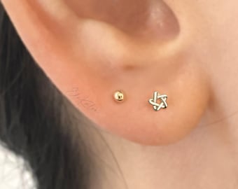 Puede incluir: Primer plano de la oreja de una persona con dos pendientes de oro. El pendiente del lóbulo inferior es un pequeño pendiente redondo. El pendiente del lóbulo superior es un pequeño pendiente en forma de estrella.