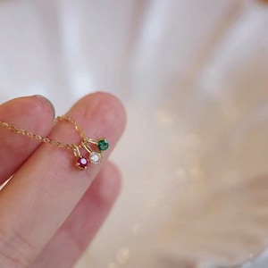 Puede incluir: Un collar de cadena de oro con tres pequeños colgantes, cada uno con una gema de color diferente: un rubí rojo, un diamante blanco y una esmeralda verde.