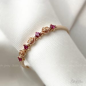 May include: A delicate gold ring with a row of small red rose accents. The ring is set on a white fabric background.