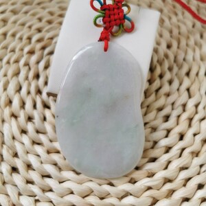 May include: A light green jade pendant with a red and white knotted cord. The pendant is shaped like a rounded oval and has a smooth, polished surface.