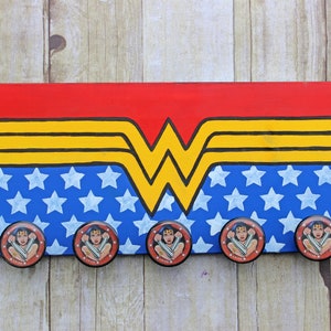 May include: A wooden sign with a Wonder Woman logo painted on it. The logo is red, yellow, and black with a blue background with white stars. There are five Wonder Woman themed knobs attached to the sign.