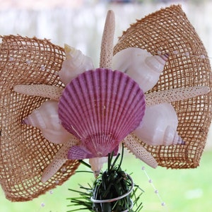 May include: A decorative tree topper featuring a burlap bow adorned with seashells, starfish, and a large purple scallop shell. White pearls and greenery add to the coastal theme. The topper is designed for a festive, beach-inspired holiday.