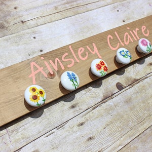 May include: Wooden name board with the name "Ainsley Claire" in pink script. The board has six white knobs with hand-painted floral designs, including sunflowers, blue flowers, and red flowers. The board is made of light brown wood.