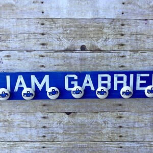 May include: A blue wooden sign with the names "Liam Gabriel" in white letters. There are six blue truck knobs attached to the sign.