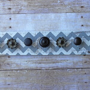 May include: A wooden wall-mounted coat rack with a chevron pattern painted in gray and white. The rack has six black metal knobs for hanging items.