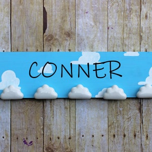 May include: A blue wooden sign with white clouds painted on it. The sign says "CONNER" in black marker. There are five white cloud-shaped hooks attached to the bottom of the sign.