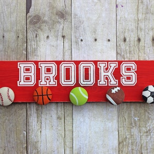 May include: A red wooden sign with the name "BROOKS" in white letters. The sign has five painted sports ball hooks: baseball, basketball, tennis ball, football, and soccer ball.