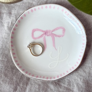 Könnte beinhalten: Eine weiße Keramik-Ringschale mit einem rosa Schleifenmuster und dem Buchstaben "L". Die Schale hat einen goldenen Rand und rosa Tupfen am Rand. Ein goldener Ring mit einem Diamanten liegt darin.