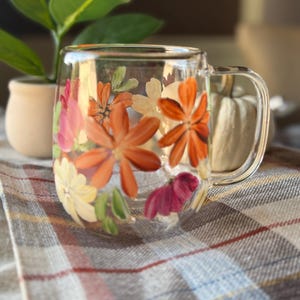 Pode incluir: Uma caneca de vidro transparente com uma alça, adornada com flores laranja, rosa e brancas pintadas à mão. O design floral é definido contra um fundo desfocado com uma planta e um cobertor xadrez.