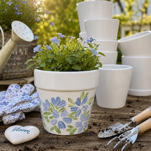 May include: A white ceramic flower pot with blue floral design, filled with green plants and blue flowers. Other white pots, gardening tools, a watering can, and a stone with the word "Bloom" are also visible, suggesting a gardening theme.