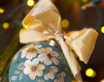 May include: A hand-painted teal Christmas ornament with white and yellow floral designs and gold accents. The ornament is tied with a gold satin bow. The background is blurred with Christmas lights.