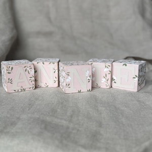May include: Wooden blocks spelling the name "ANN H". Each block is painted light pink with white floral accents and a letter in a light beige color. The blocks are arranged on a neutral-colored surface.