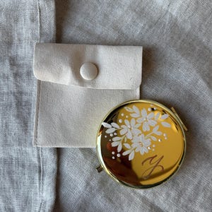 May include: A gold compact mirror with a floral design and the letter "Y". The mirror is next to a small, light-colored pouch with a button closure. The background is a light gray fabric.