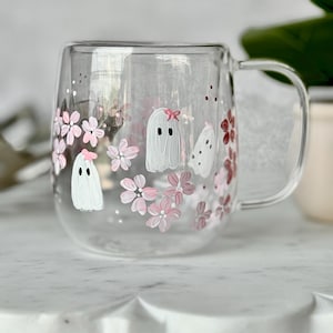 May include: A clear glass mug with a handle, decorated with hand-painted pink cherry blossoms and three white ghost figures. One ghost wears a pink bow. The mug is on a white surface.