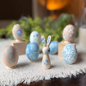 May include: A collection of Easter decorations featuring wooden eggs and a bunny figurine. The eggs are painted in light blue and beige with floral patterns. The bunny is light brown with blue floral accents, all arranged on a white cloth.