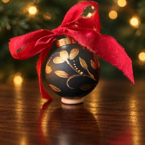 May include: A spherical matte black Christmas ornament featuring hand-painted gold floral motifs and a metallic gold cap. A vibrant red fabric ribbon is tied in a bow at the top of the ornament, which rests on a small, light-colored wooden base.