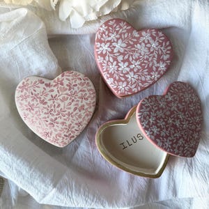 May include: Three heart-shaped trinket boxes. Two are closed, and one is open, revealing the word "ILUS" inside. The boxes are pink with white floral patterns and gold trim. They rest on a white, textured cloth.