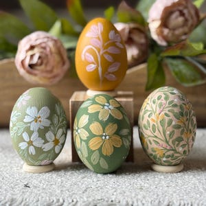 May include: Hand-painted decorative Easter eggs in various colors, including mustard yellow, olive green, and peach. Each egg features a unique floral design in white, pink, and yellow. The eggs are displayed on small wooden stands.