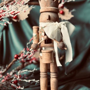 Puede incluir: Una figura de cascanueces de madera con una corona, sosteniendo baquetas y un pequeño tambor. La figura está pintada con detalles florales y tiene una cinta blanca alrededor del cuello. Bayas rojas y ramas en el fondo.