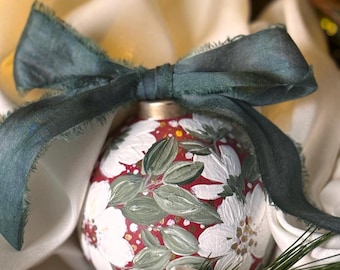 May include: A hand-painted Christmas ornament with a red background and white floral design, accented with green leaves. The ornament is tied with a dark green ribbon. The background is a soft, off-white fabric.