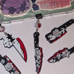 May include: A phone case with stickers, including a jar of books and a ramen bowl. Hanging from the case are black and red knife charms with floral and bunny designs. The text "Bookish Bar" is visible.