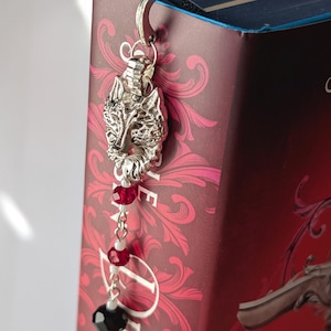 May include: Silver wolf head bookmark with red and black beads. The bookmark is attached to a book with a burgundy cover and floral pattern. The bookmark features a wolf head, beads, and a crescent moon charm.