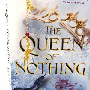 Puede incluir: Un marcap&aacute;ginas de metal dorado con un dije de serpiente y una cuenta roja cuelga de la parte superior de un libro titulado "The Queen of Nothing" de Holly Black. La portada del libro presenta una ilustraci&oacute;n de serpiente y letras doradas.