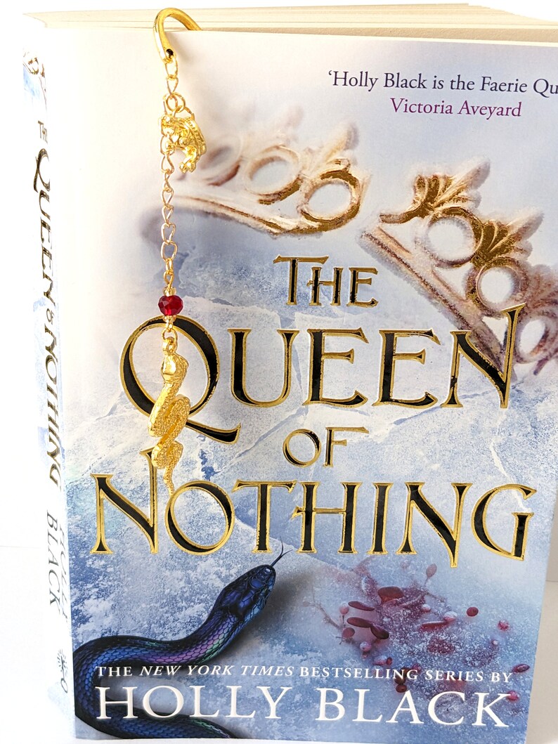 Puede incluir: Un marcap&aacute;ginas de metal dorado con un dije de serpiente y una cuenta roja cuelga de la parte superior de un libro titulado "The Queen of Nothing" de Holly Black. La portada del libro presenta una ilustraci&oacute;n de serpiente y letras doradas.