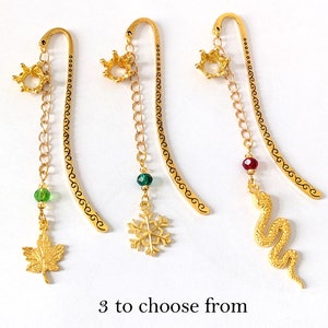 Puede incluir: Tres marcap&aacute;ginas de metal dorado con abalorios decorativos. Cada marcap&aacute;ginas presenta una corona en la parte superior, una cadena y un abalorio: una hoja de arce, un copo de nieve y una serpiente. El texto "3 to choose from" est&aacute; en la parte inferior.