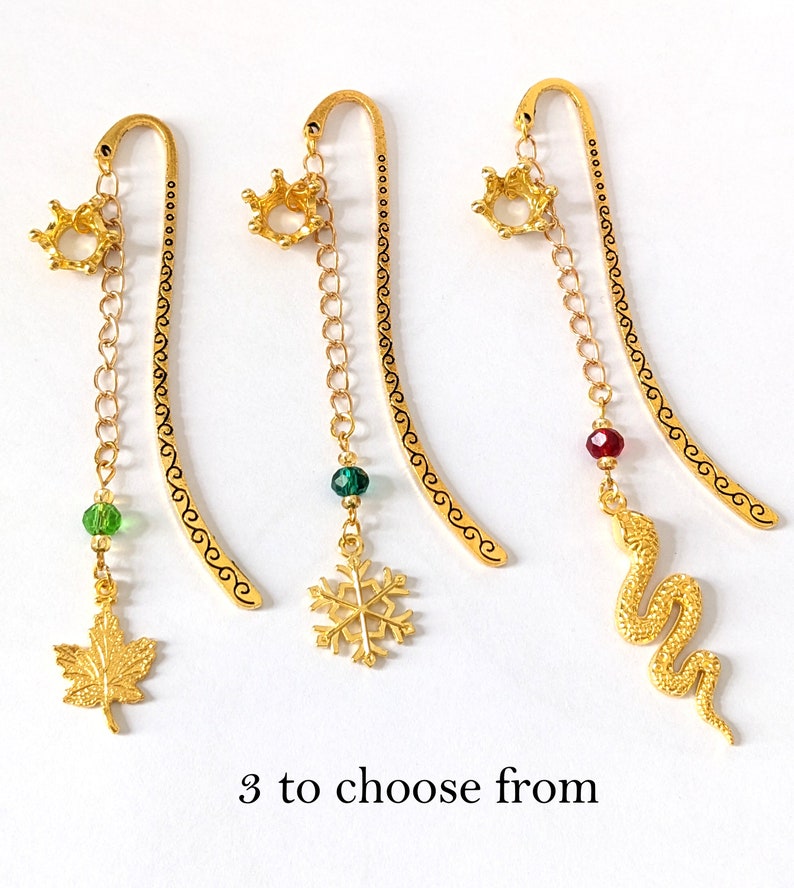 Puede incluir: Tres marcap&aacute;ginas de metal dorado con abalorios decorativos. Cada marcap&aacute;ginas presenta una corona en la parte superior, una cadena y un abalorio: una hoja de arce, un copo de nieve y una serpiente. El texto "3 to choose from" est&aacute; en la parte inferior.