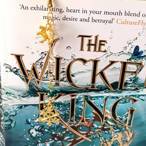Puede incluir: Un marcap&aacute;ginas dorado con un dije de corona y copo de nieve est&aacute; sujeto a un libro titulado "The Wicked King". El marcap&aacute;ginas cuelga de la parte superior del libro. La portada del libro tiene un dise&ntilde;o en color turquesa y dorado.