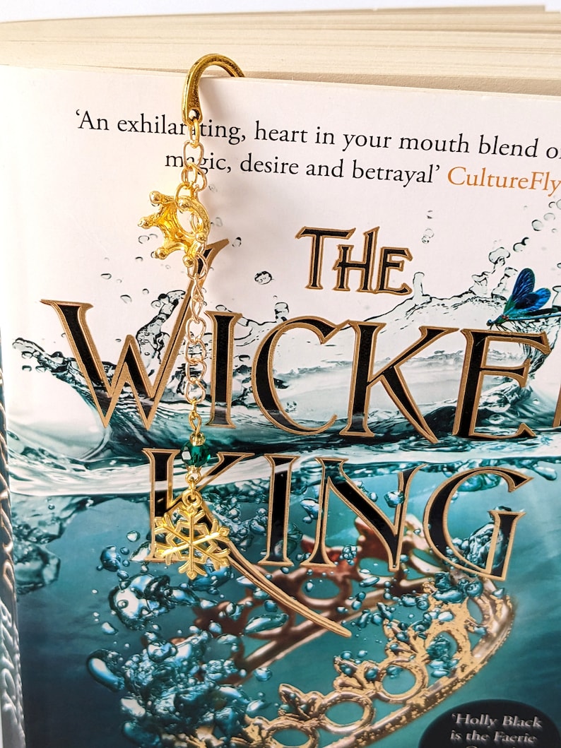 Puede incluir: Un marcap&aacute;ginas dorado con un dije de corona y copo de nieve est&aacute; sujeto a un libro titulado "The Wicked King". El marcap&aacute;ginas cuelga de la parte superior del libro. La portada del libro tiene un dise&ntilde;o en color turquesa y dorado.