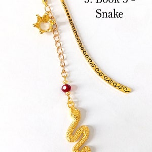Puede incluir: Un marcap&aacute;ginas dorado con un dije de serpiente. El marcap&aacute;ginas tiene un dije de corona y una cuenta roja. El texto "3. Book 3 - Snake" es visible en la imagen.