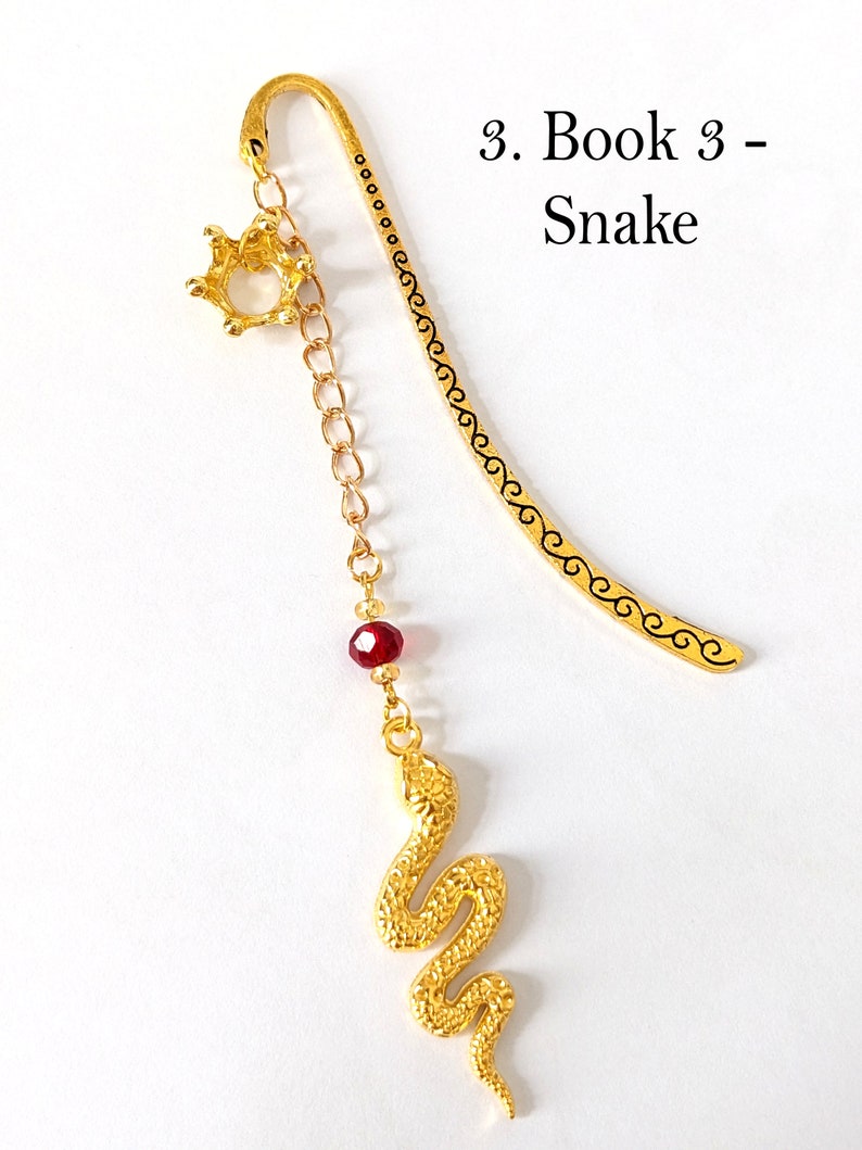 Puede incluir: Un marcap&aacute;ginas dorado con un dije de serpiente. El marcap&aacute;ginas tiene un dije de corona y una cuenta roja. El texto "3. Book 3 - Snake" es visible en la imagen.