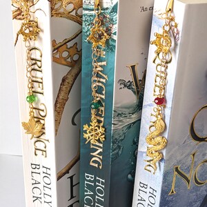 Puede incluir: Marcadores de metal dorado con dijes decorativos cuelgan de la parte superior de tres libros. Los dijes incluyen una hoja de arce, un copo de nieve y una serpiente. Las lomos de los libros muestran los t&iacute;tulos "Cruel Prince", "Wicked King" y "Queen of Nothing".