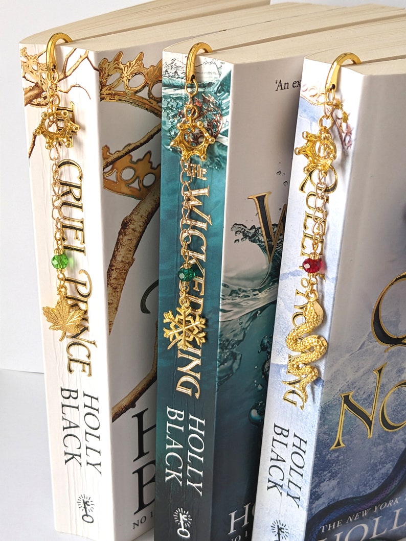Puede incluir: Marcadores de metal dorado con dijes decorativos cuelgan de la parte superior de tres libros. Los dijes incluyen una hoja de arce, un copo de nieve y una serpiente. Las lomos de los libros muestran los t&iacute;tulos "Cruel Prince", "Wicked King" y "Queen of Nothing".