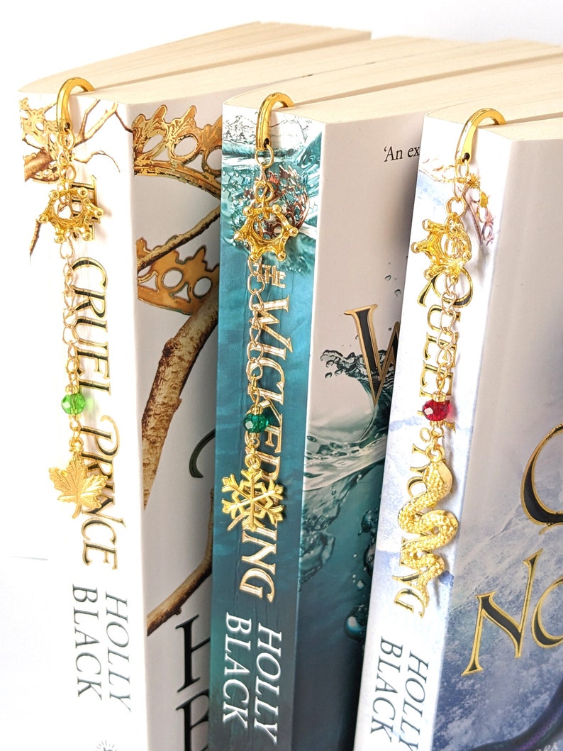 Puede incluir: Marcadores de metal dorado con colgantes insertados en las p&aacute;ginas de los libros. Los colgantes incluyen una hoja de arce, un copo de nieve y una serpiente. Los t&iacute;tulos de los libros incluyen "Cruel Prince", "Wicked King" y "Queen of Nothing".
