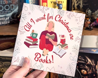 May include: A square card with the text "All I want for Christmas are Books!" in red script. The illustration shows a person reading, surrounded by books, a mug, and festive decorations. The card is held in a hand.