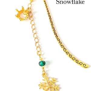 Puede incluir: Marcap&aacute;ginas de metal dorado con un dije de corona, una cuenta verde y un dije de copo de nieve. El marcap&aacute;ginas tiene una parte superior curvada y un dise&ntilde;o decorativo a lo largo del borde. El texto en la imagen dice "2. Libro 2 - Copo de nieve."