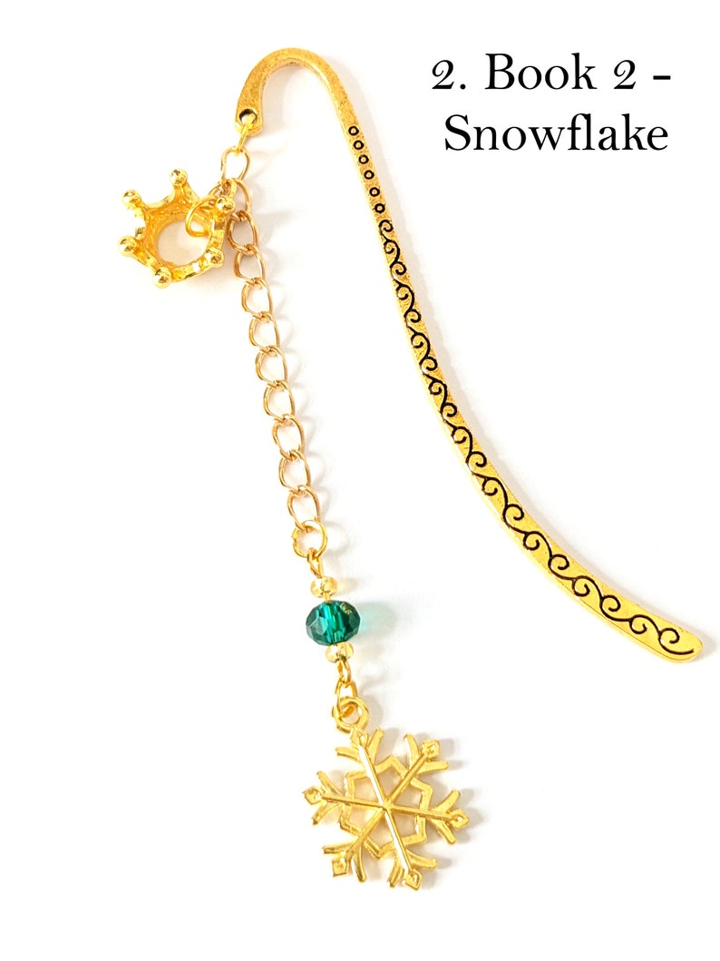 Puede incluir: Marcap&aacute;ginas de metal dorado con un dije de corona, una cuenta verde y un dije de copo de nieve. El marcap&aacute;ginas tiene una parte superior curvada y un dise&ntilde;o decorativo a lo largo del borde. El texto en la imagen dice "2. Libro 2 - Copo de nieve."