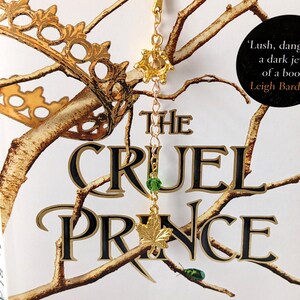 Puede incluir: Marcap&aacute;ginas dorado con una parte superior en forma de corona y una cadena que sostiene una cuenta verde y un dije de hoja de arce. El marcap&aacute;ginas est&aacute; en un libro titulado "The Cruel Prince" de Holly Black.