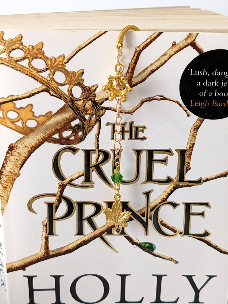 Puede incluir: Marcap&aacute;ginas dorado con una parte superior en forma de corona y una cadena que sostiene una cuenta verde y un dije de hoja de arce. El marcap&aacute;ginas est&aacute; en un libro titulado "The Cruel Prince" de Holly Black.