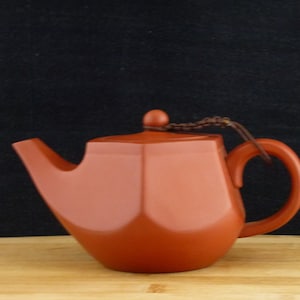 Könnte beinhalten: Eine Terrakotta-Teekanne mit einem braunen Schnursteifen als Henkel. Die Teekanne hat eine geometrische Form mit einem abgerundeten Boden und einem flachen Deckel.