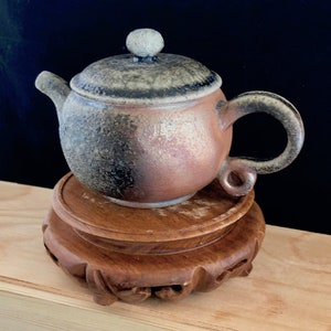 May include: A small, round, brown and grey ceramic teapot with a lid and a handle. The teapot is sitting on a wooden stand.