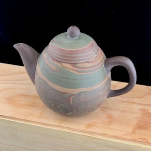 May include: A brown teapot with a green, brown, and orange marbled glaze. The teapot has a round body and a small spout. The teapot is sitting on a wooden surface.